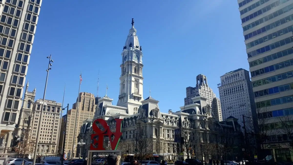 Installation emblématique "LOVE" en lettres rouges dans Love Park, avec l'Independence Hall visible en arrière-plan sous un ciel bleu. La scène symbolise l'amour et l'histoire de Philadelphie
