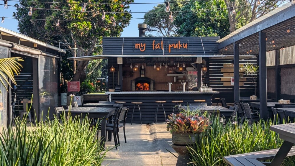 Photo du café "My Fat Puku" sur la Grande Barrière de Nouvelle-Zélande, avec des lettres orange sur une ardoise noire et un four à pizza