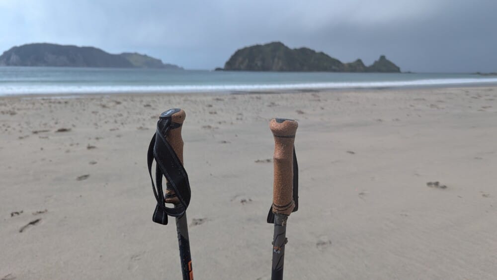 Photo d'un paysage de plage sur la Grande Barrière de Nouvelle-Zélande, avec un bâton de randonnée au premier plan et des rochers au loin