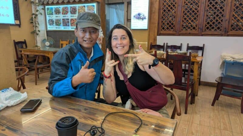 Inès assise avec un serveur dans un restaurant à Dali, Chine, tous deux souriants et faisant un signe cool avec les mains