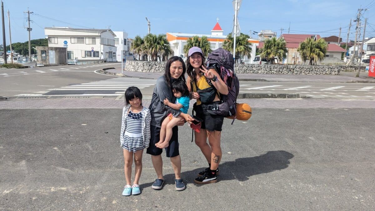 Inès souriante avec une mère japonaise et ses deux filles sur le parking à Yoron Island, Japon. Moment convivial et joyeux lors d’un autostop, ambiance chaleureuse et humaine