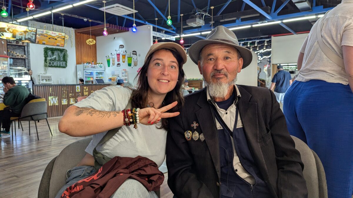 Inès en casquette fait un signe peace à Oulan-Bator en Mongolie. Elle pose aux côtés d’un vieux monsieur mongol dans un portrait urbain
