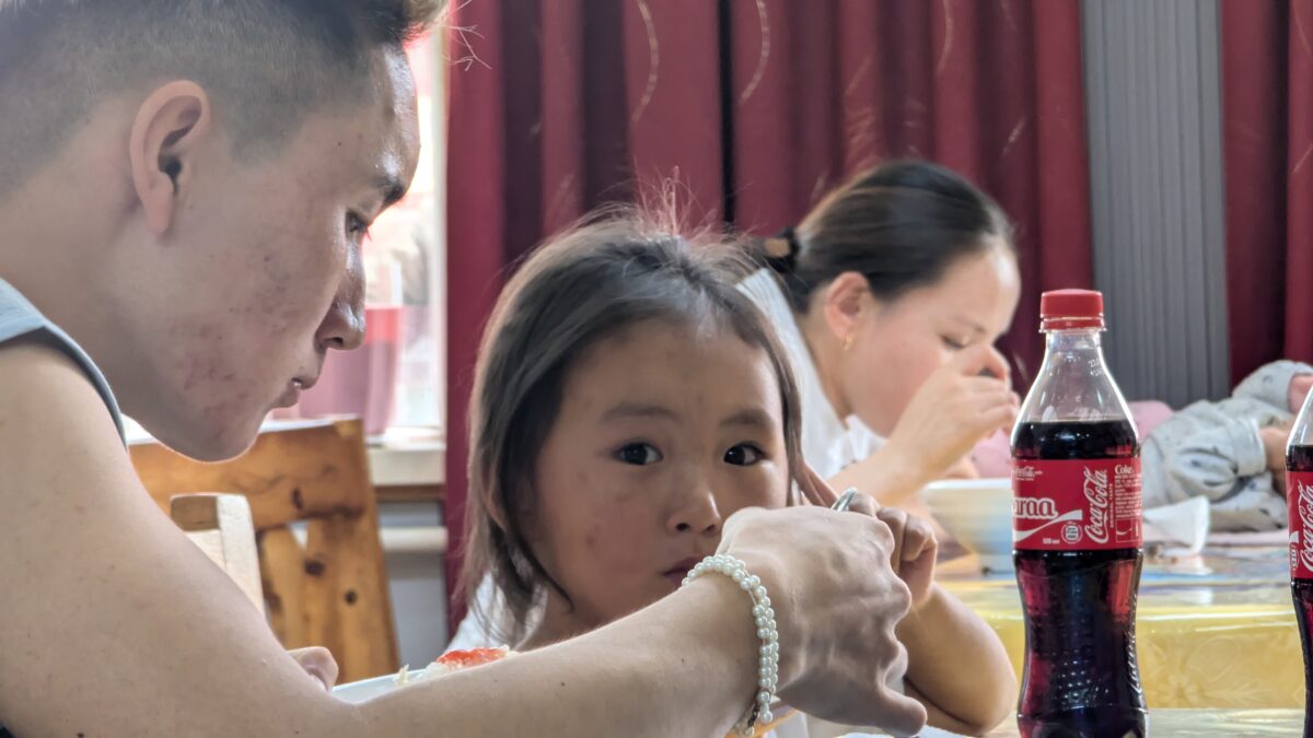 Portrait d’un jeune nomade au premier plan en Mongolie, dans un intérieur de vie quotidienne. En arrière-plan, une petite fille assise à une table partage un moment de vie simple
