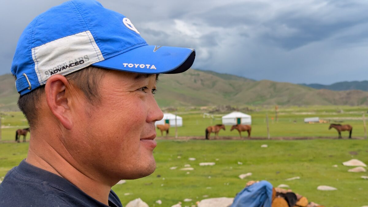 Portrait d’un homme mongol en casquette, de profil, observant la steppe. À l’arrière-plan : chevaux, yourte traditionnelle et vastes plaines de Mongolie