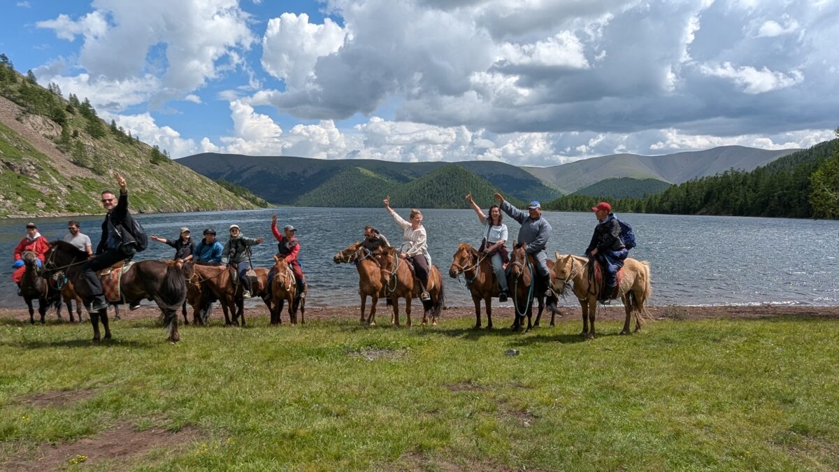 Groupe de voyageurs et nomades avec Inès levant les bras devant un lac en Mongolie. Chevaux présents dans un paysage de la vallée de l’Orkhon