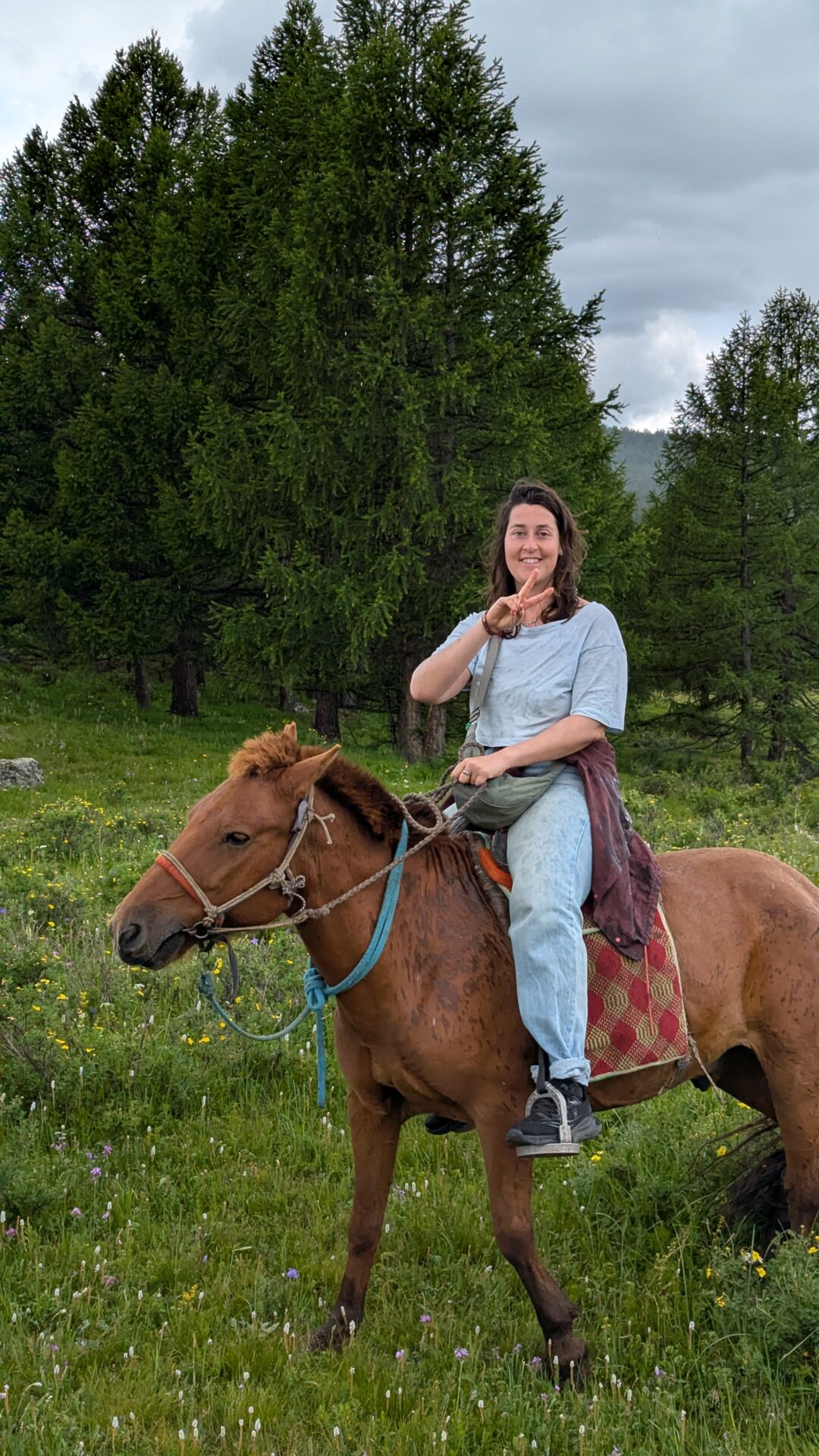 Inès à cheval dans la vallée de l’Orkhon en Mongolie, faisant un signe peace. Scène de chevauchée en pleine steppe