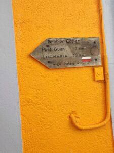 Indications en bois sur un mur jaune pour le sentier côtier GR340 au Palais, Belle-Ile-en-Mer, France, avec le symbole du GR