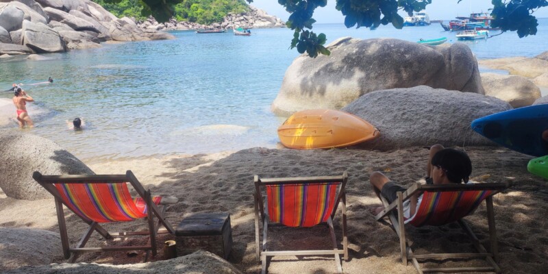 Transats rouges alignés sur la plage de Koh Tao, avec un bateau en arrière-plan et des eaux bleu turquoise