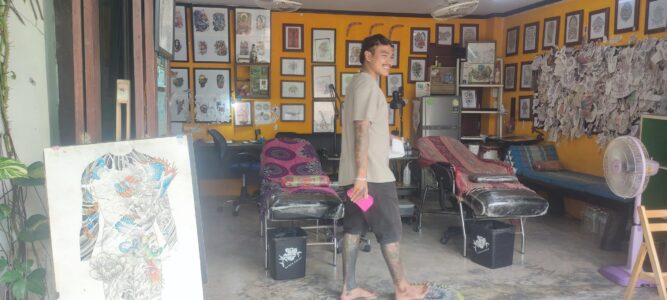 Un homme souriant au centre d'un salon de tatouage extérieur à Koh Tao, entouré de décorations artistiques et d'équipements de tatouage