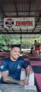 Un local souriant avec un tatouage, posé derrière un ring de boxe à Koh Tao, dans une ambiance d'entraînement dynamique