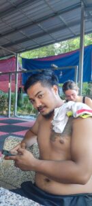 Un boxeur thaïlandais torse nu, assis et souriant à Koh Tao, entouré de sacs de frappe et d'équipements de Muay Thai