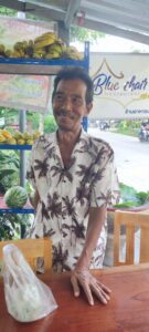 Un marchand local souriant portant une chemise à fleurs rose à Koh Tao, debout devant son étal