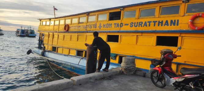 Un homme attend avec son scooter devant un bateau de transport jaune à Koh Tao