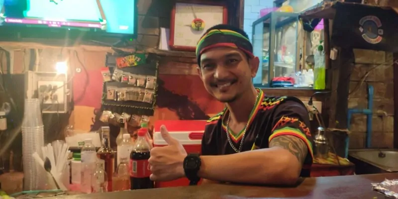 Un homme rasta souriant faisant un signe de paix à l'intérieur d'un bar rasta à Koh Tao, entouré de décorations colorées et reggae