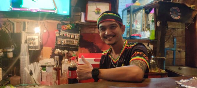 Un homme rasta souriant faisant un signe de paix à l'intérieur d'un bar rasta à Koh Tao, entouré de décorations colorées et reggae