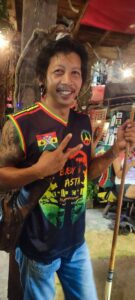 Un homme rasta souriant faisant un signe de paix à l'intérieur d'un bar rasta à Koh Tao, entouré de décorations colorées et reggae