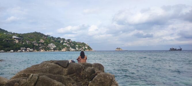 Photographie d'une femme de dos sur des rochers à Koh Tao, avec une baie et un rocher en arrière-plan