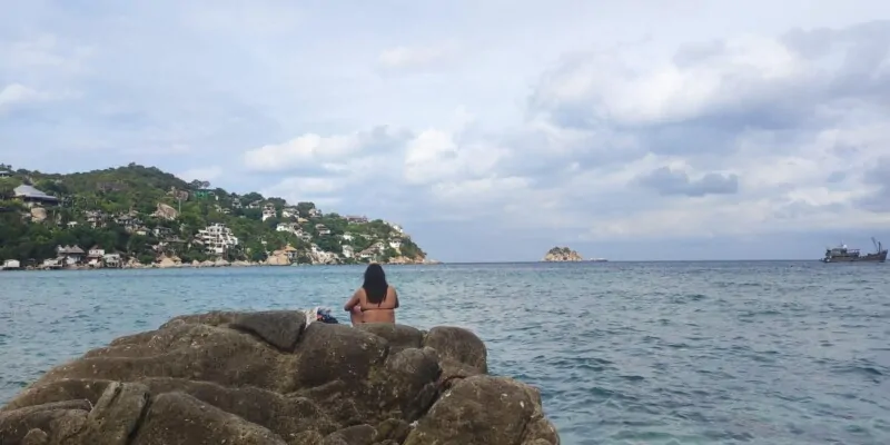 Photographie d'une femme de dos sur des rochers à Koh Tao, avec une baie et un rocher en arrière-plan