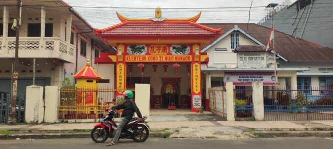 Photographie d'un homme en scooter, portant un casque vert, s'arrêtant devant un temple chinois à Manado