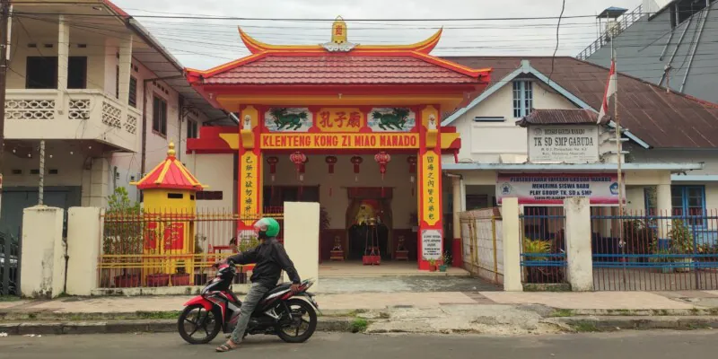 Photographie d'un homme en scooter, portant un casque vert, s'arrêtant devant un temple chinois à Manado