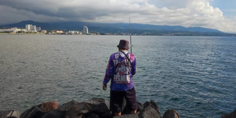 Portrait d'un homme de dos, tenant une canne à pêche, avec des immeubles et des montagnes de Manado en arrière-plan