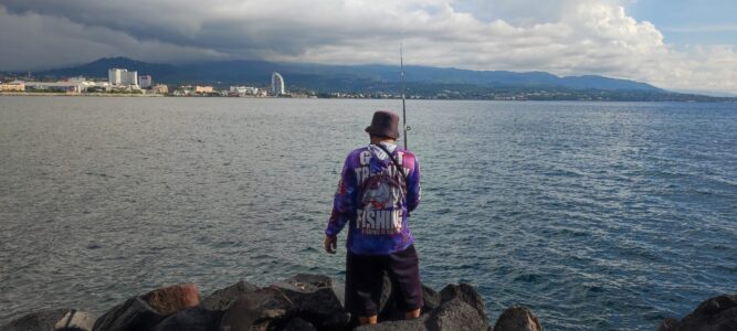 Portrait d'un homme de dos, tenant une canne à pêche, avec des immeubles et des montagnes de Manado en arrière-plan