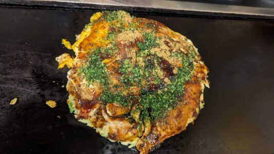 Okonomiyaki : pancakes japonais Gros plan d'un Okonomiyaki, un plat japonais, garni de mayonnaise, de sauce takoyaki, de flocons de bonite et d'oignons verts