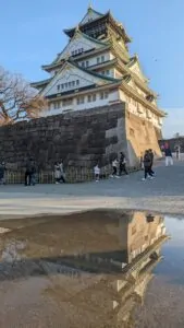 Image du château d'Osaka se reflétant dans l'eau, avec son architecture traditionnelle entourée de végétation, créant une scène harmonieuse et paisible