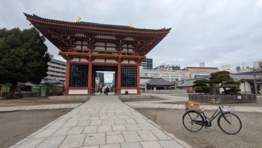 La scène montre l’entrée du Shitennō-ji, l'un des plus anciens temples bouddhistes du Japon, situé à Osaka. Des vélos sont garés à l’extérieur, symbolisant la manière dont les habitants intègrent la culture cycliste dans leur quotidien. Le portail majestueux du temple, en bois sombre avec des toits à pignons, se dresse en arrière-plan, orné de sculptures et de détails architecturaux typiques de l’époque Asuka. Les visiteurs, certains en tenues traditionnelles, entrent et sortent du temple, ajoutant une touche vivante à l’ambiance. Cette image capture l’harmonie entre le patrimoine spirituel ancien et la vie quotidienne moderne dans une grande ville japonaise