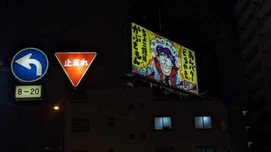 Affiche de "Chercher Charlie" accrochée dans la rue d'Osaka, entourée de panneaux de directions, dans une ambiance néo-grunge nocturne