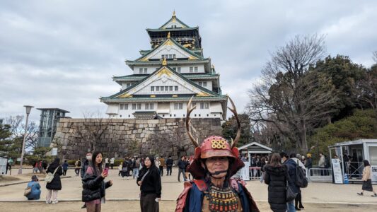 Homme en costume traditionnel japonais avec un casque à cornes de cerf, souriant devant le Château d'Osaka