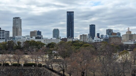 Vue panoramique depuis le château d'Osaka, montrant la ville moderne entourée de zones verdoyantes, avec des arbres et des monuments historiques en premier plan, et des gratte-ciels en arrière-plan