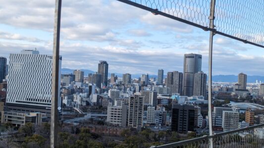 Vue de la ville d’Osaka à travers un grillage depuis le château d'Osaka, avec des gratte-ciels à l'horizon et des jardins verdoyants au premier plan