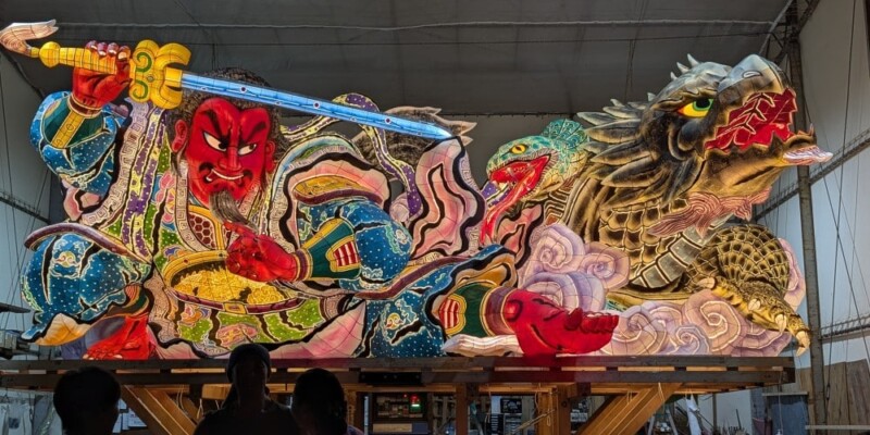 "Photographie de chars Nebuta à l'arrêt pendant le festival, ornés de décorations colorées et illuminés, avec des participants en costumes traditionnels autour