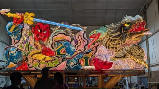 "Photographie de chars Nebuta à l'arrêt pendant le festival, ornés de décorations colorées et illuminés, avec des participants en costumes traditionnels autour