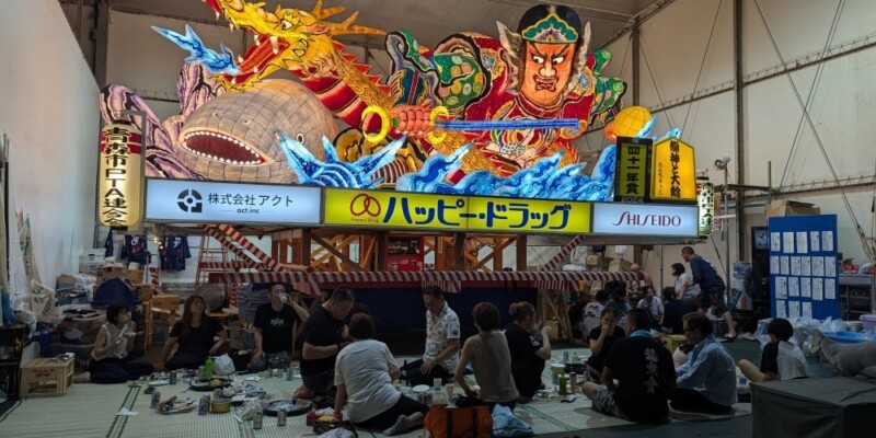 "Photographie de chars Nebuta à l'arrêt pendant le festival, ornés de décorations colorées et illuminés, avec des participants en costumes traditionnels se relaxant et mangeant en dessous