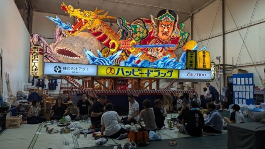 "Photographie de chars Nebuta à l'arrêt pendant le festival, ornés de décorations colorées et illuminés, avec des participants en costumes traditionnels se relaxant et mangeant en dessous