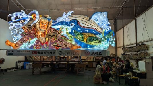 "Photographie de chars Nebuta à l'arrêt pendant le festival, ornés de décorations colorées et illuminés, avec des participants en costumes traditionnels se relaxant et mangeant en dessous