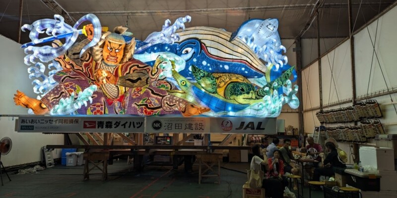 "Photographie de chars Nebuta à l'arrêt pendant le festival, ornés de décorations colorées et illuminés, avec des participants en costumes traditionnels se relaxant et mangeant en dessous