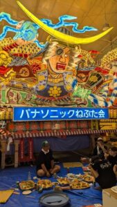 "Photographie de chars Nebuta à l'arrêt pendant le festival, ornés de décorations colorées et illuminés, avec des participants en costumes traditionnels se relaxant et mangeant en dessous