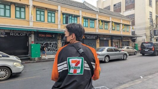 Portrait d'un homme de dos, vêtu d'un t-shirt et d'un short, se tenant devant un magasin 7-Eleven sur Khao San Road à Bangkok, avec des néons colorés et une foule animée en arrière-plan