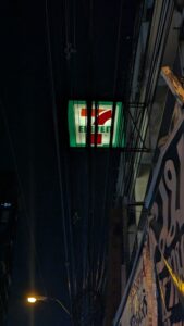 Vue nocturne d'un magasin 7-Eleven à Bangkok, illuminé par des néons, avec un enchevêtrement de fils électriques visibles au-dessus du bâtiment