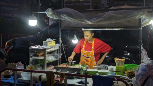 Stand de street food illuminé par des ampoules, avec une femme en train de cuisiner sous la lumière nocturne de Bangkok