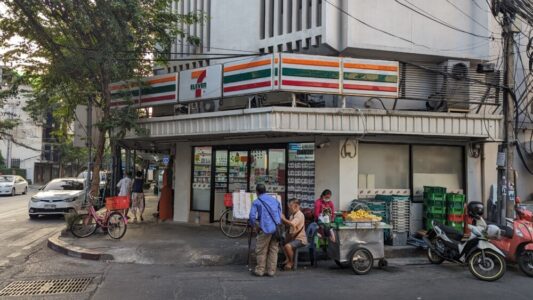Un 7-Eleven à Bangkok avec plusieurs personnes devant, à côté d'un stand de street food