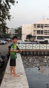Portrait d'un enfant local souriant, tenant une canne à pêche, portant un t-shirt vert au bord du fleuve à Bangkok