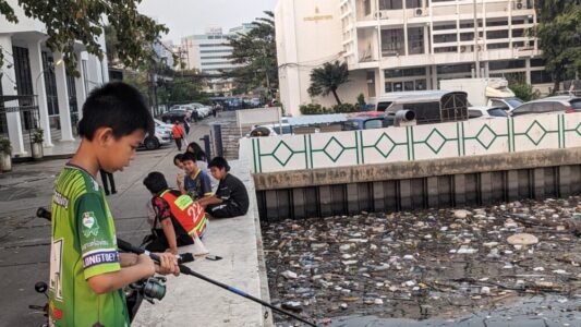 Portrait d'un enfant local souriant, tenant une canne à pêche, portant un t-shirt vert au bord du fleuve à Bangkok