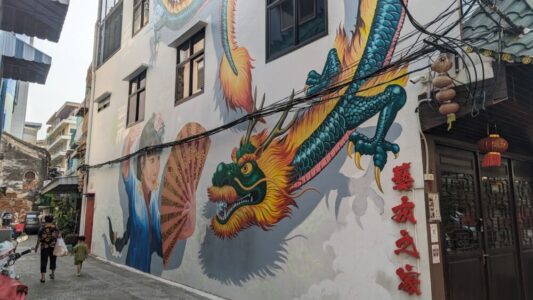 Illustration de street art représentant un dragon entier en jaune et vert avec une moustache, sur un mur, à côté d'une petite fille chinoise
