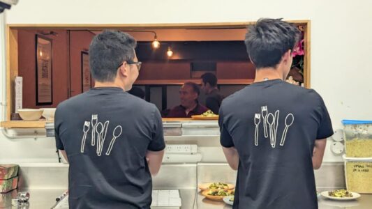 Deux volontaires de dos à Wellington, en tee-shirts noirs avec des couverts imprimés. Ils assurent le service lors d’un événement